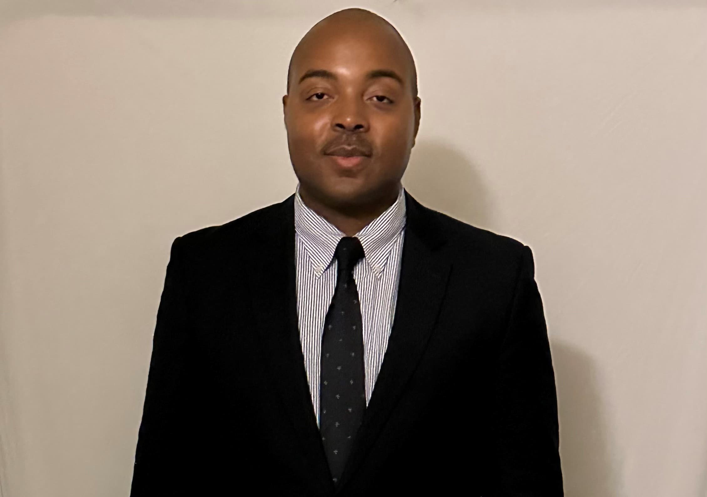 Bald Black man in a black suit, striped shirt, and dark patterned tie.